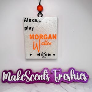 May include: A rectangular, glittery white air freshener with the text "Alexa... play MORGAN Wallen" in black and orange. The design includes music player icons. A purple and white "MakeScents Freshies" logo is below.