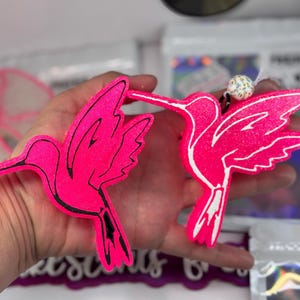 May include: Two vibrant pink hummingbird ornaments with black outlines and white accents. The hummingbirds are held in a hand, showcasing their detailed design. Each ornament has a small, decorative bead attached.