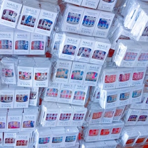 Puede incluir: Una gran variedad de uñas artificiales empaquetadas. Las cajas blancas llevan la palabra "NAILS" impresa. Los diseños de uñas varían en color y estilo, incluyendo purpurina, patrones y colores lisos.