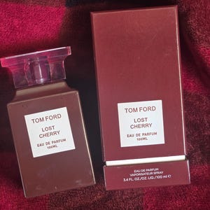 Op de afbeelding: Een bruine rechthoekige parfumfles en bijpassende doos. De fles heeft een transparante, rechthoekige dop. Zowel de fles als de doos hebben een wit etiket met de tekst "TOM FORD LOST CHERRY EAU DE PARFUM 100ML". De doos bevat ook de tekst "3.4 FL. OZ./OZ. LIQ./100 ml e".