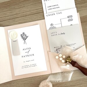 以下が含まれることがあります： ライトピンクのフォルダーに入った結婚式の招待状セット。花柄のイラスト、AlisaとPatrickの名前、23. August 2026の日付が記載された白いカードが含まれています。詳細カード、タイムラインカード、ワックスシールスタンプ、白いワックスビーズがセットになっています。