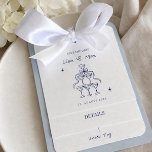 Può includere: Un biglietto d'invito di nozze bianco e azzurro con un fiocco di nastro bianco. Il biglietto recita "Save the Date" e include i nomi "Lisa & Max" e la data "23 agosto 2026". È presente anche una grafica di calici di champagne.