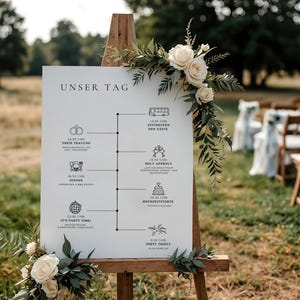 May include: A white sign with a wooden easel, decorated with white flowers and greenery, lists the schedule for a wedding day. The sign reads "Unser Tag" at the top and includes details like "Free Traubing", "Holy & Sperrly", "Dinner", "It's Party Time", and "Party Start".