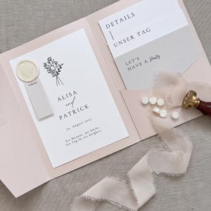 以下が含まれることがあります： ペールピンクのウェディング招待状フォルダー。中にはAlisaとPatrickの名前、花のイラスト、23. August 2027の日付が記された白いカードが入っています。ワックスシール、詳細カード、パーティーカード、ほつれたライトピンクのリボンが添えられています。
