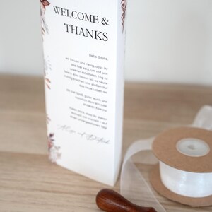 May include: A white wedding program with the words "WELCOME & THANKS" in black. Floral accents in brown and burgundy decorate the edges. A wooden wax seal, white ribbon, and a spool of ribbon are also visible.