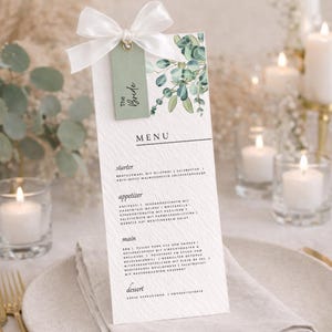 May include: A wedding menu card with a white ribbon and a green tag that says "The Bride". The menu features a watercolor eucalyptus design. The menu is on a white plate with a linen napkin and gold silverware.