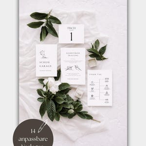 May include: A collection of white wedding stationery cards featuring black text and line art illustrations, including a table number, a shoe garage sign, a ceremony program, and a timeline card, accented by green leaves and a small ring box.