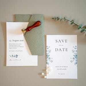 Könnte beinhalten: Hochzeitspapeterie-Set mit einer 'Save the Date'-Karte und einer Einladung. Die Einladung enthält das Datum '23. August 2026'. Blaue florale Akzente verzieren die Karten. Ein Wachssiegel mit Holzgriff und ein grüner Umschlag sind ebenfalls vorhanden.