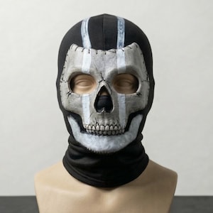 Puede incluir: Un pasamontañas negro con un diseño de calavera. La calavera es principalmente gris y blanca, con cuencas de ojos y nariz negras. Rayas blancas verticales recorren la parte superior de la cabeza. El pasamontañas se muestra en una cabeza de maniquí.