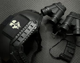 Könnte beinhalten: Schwarzer taktischer Helm mit Totenkopf-Patch und Nachtsichtbrille. Die Brille ist an einer Halterung befestigt und mit einem Akku verbunden. Alle Komponenten sind schwarz.