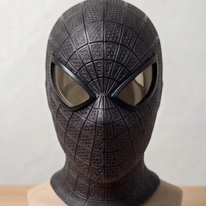 May include: A full-head mask featuring a dark charcoal gray textured surface with a raised grid pattern and hexagonal detailing. The mask includes two large, almond-shaped eye openings covered with a fine, metallic gold-toned mesh screen.
