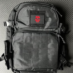 Puede incluir: Mochila táctica negra con un parche de calavera roja en la parte delantera. La mochila tiene varios bolsillos y correas.