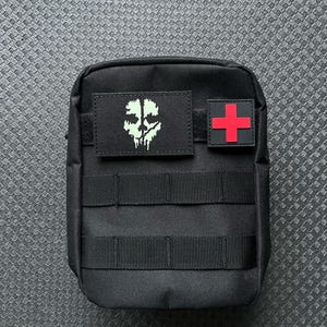 Könnte beinhalten: Schwarze taktische Tasche mit einem grünen Totenkopf- und Knochen-Patch und einem roten medizinischen Kreuz-Patch.