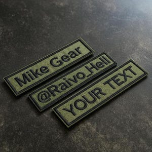 Op de afbeelding: Drie rechthoekige olijfgroene patches met zwart geborduurde tekst. De bovenste patch leest "Mike Gear", de middelste patch leest "@Raivo_Hell" en de onderste patch leest "YOUR TEXT". De patches hebben een zwarte rand.