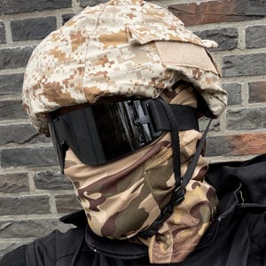 Op de afbeelding: Een helm met een bruin en bruin camouflagepatroon, een zwarte bril en een bijpassende nekwarmer. De persoon draagt een zwarte jas. De items lijken militaire of tactische uitrusting te zijn.