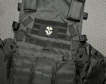 Könnte beinhalten: Schwarze taktische Weste mit mehreren Taschen und einem Totenkopf-Patch. Die Weste hat verstellbare Träger und einen Reißverschluss.