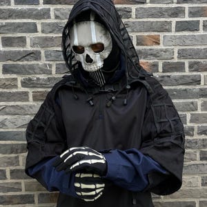 May include: A person wearing a black hooded jacket with a skull mask and skeleton gloves. The jacket has a mesh hood and a zipper closure. The person is standing in front of a brick wall.