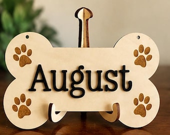 Könnte beinhalten: Ein helles Holzschild in Form eines Hundeknochens mit dem Namen August in erhabener schwarzer Schrift. Vier braune Pfotenabdrücke sind in die Ecken des Knochens eingraviert. Das Schild steht auf einem kleinen Holzständer.