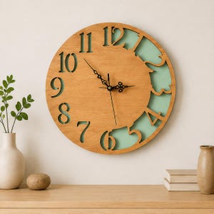 May include: A round wall clock featuring a light wood grain face with large, cut-out numerals from 4 to 12. The background behind the cut-outs is a solid pale mint green. The clock has black metal hour, minute, and second hands at the centre.