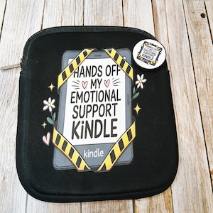 May include: Black Kindle case with a zip and a button. The case has a graphic that reads "HANDS OFF MY EMOTIONAL SUPPORT KINDLE" with yellow and black hazard tape design. The button has the same text.