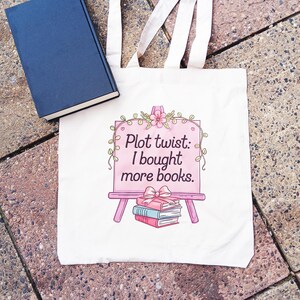May include: A white tote bag with the text "Plot twist: I bought more books" in a pink frame with a stack of books. A blue book rests on the side. The bag has two white straps.