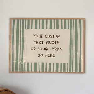 Puede incluir: Un cuadro rectangular en un marco de madera clara con un panel central color crema y el texto en marrón oscuro: YOUR CUSTOM TEXT, QUOTE OR SONG LYRICS GO HERE. El borde presenta rayas verticales de color verde salvia sobre un fondo crema.