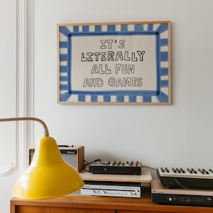 Pu&ograve; includere: Una stampa artistica da parete rettangolare in una cornice di legno chiaro con un bordo a righe blu dipinto a mano su sfondo crema. Il testo al centro recita in lettere maiuscole nere: IT'S LITERALLY ALL FUN AND GAMES. La stampa &egrave; appesa a una parete.