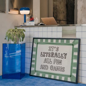 May include: A rectangular framed print featuring a white background with a green painted border and the black text IT'S LITERALLY ALL FUN AND GAMES. The artwork is positioned on the floor next to a blue acrylic pedestal and a white tiled counter.