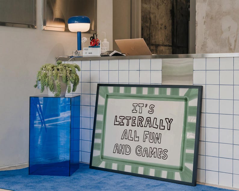May include: A rectangular framed print featuring a white background with a green painted border and the black text IT'S LITERALLY ALL FUN AND GAMES. The artwork is positioned on the floor next to a blue acrylic pedestal and a white tiled counter.