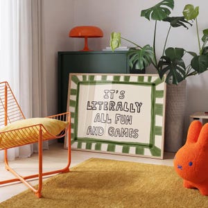 May include: A rectangular framed wall art piece featuring a cream-colored background with a hand-painted green striped border. The center text in black, hand-drawn style lettering reads: IT'S LITERALLY ALL FUN AND GAMES, enclosed in a light wood frame.