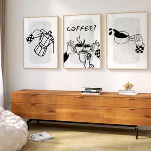 May include: Three framed art prints with coffee-themed illustrations. Each print features black and white line drawings of hands interacting with coffee-making tools and cups. The middle print has the word "COFFEE?" in bold letters. The frames are gold-colored.