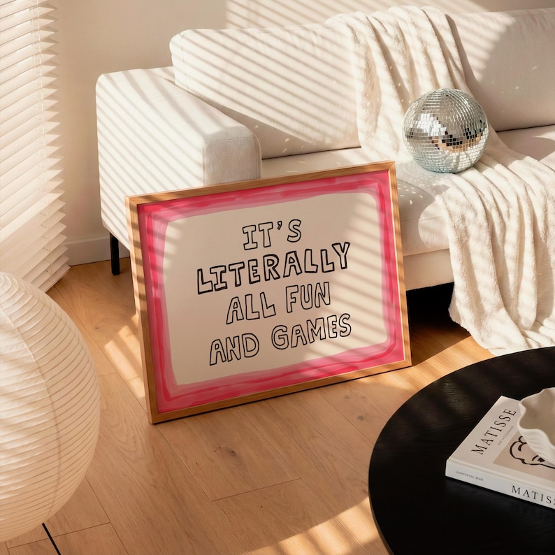 Pu&ograve; includere: Un'opera d'arte da parete rettangolare incorniciata con sfondo bianco, un bordo dipinto in rosa e il testo nero "IT'S LITERALLY ALL FUN AND GAMES" centrato al centro. La cornice &egrave; in legno chiaro e poggia sul pavimento.