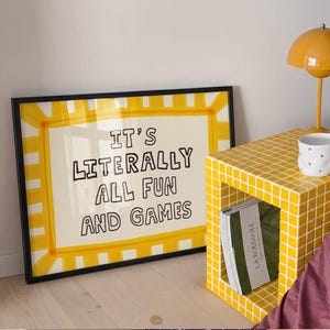 May include: A rectangular framed print with a yellow and white striped border and the black text IT'S LITERALLY ALL FUN AND GAMES. The print is positioned next to a yellow tiled cube side table, a white mug with small red hearts, and a yellow lamp.