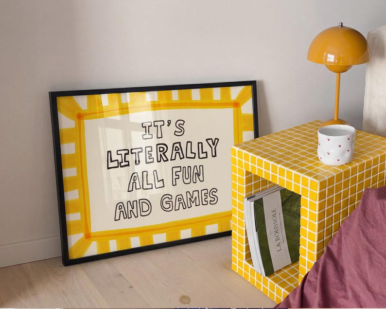 May include: A rectangular framed print with a yellow and white striped border and the black text IT'S LITERALLY ALL FUN AND GAMES. The print is positioned next to a yellow tiled cube side table, a white mug with small red hearts, and a yellow lamp.
