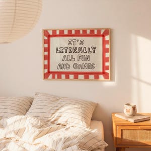 May include: A rectangular wall art print in a light wood frame featuring a red and white striped border. The center contains the black hand-lettered text IT'S LITERALLY ALL FUN AND GAMES on a cream background, displayed above a bed with striped bedding.