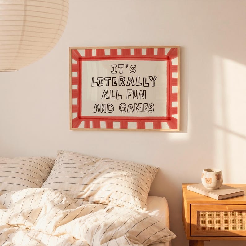 May include: A rectangular wall art print in a light wood frame featuring a red and white striped border. The center contains the black hand-lettered text IT'S LITERALLY ALL FUN AND GAMES on a cream background, displayed above a bed with striped bedding.