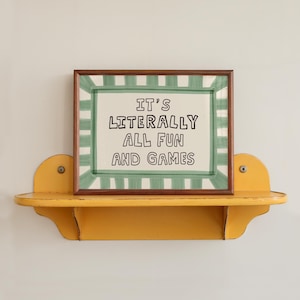 Pu&ograve; includere: Una stampa rettangolare incorniciata con sfondo bianco e un bordo a righe verdi. Il testo al centro recita "IT'S LITERALLY ALL FUN AND GAMES" in lettere maiuscole nere. La cornice &egrave; marrone scuro e poggia su una mensola da parete gialla.