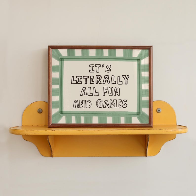 Pu&ograve; includere: Una stampa rettangolare incorniciata con sfondo bianco e un bordo a righe verdi. Il testo al centro recita "IT'S LITERALLY ALL FUN AND GAMES" in lettere maiuscole nere. La cornice &egrave; marrone scuro e poggia su una mensola da parete gialla.