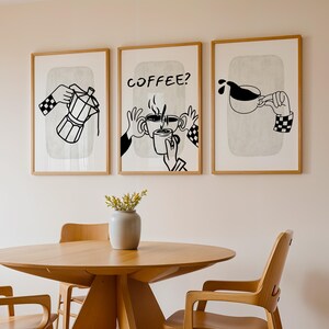 May include: Three framed art prints with coffee-themed illustrations. The prints feature black and white line drawings of coffee pots, hands holding mugs, and pouring coffee. The frames are light brown, and the prints hang above a wooden table and chairs.