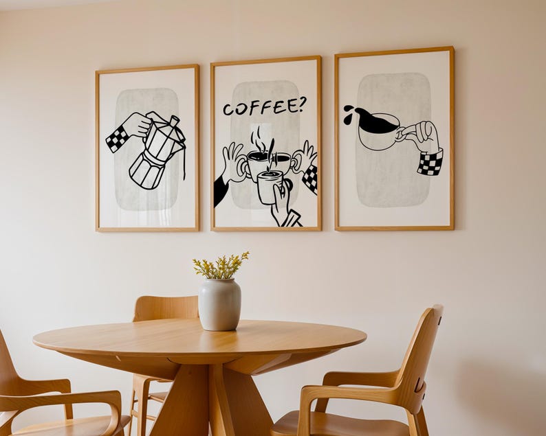 May include: Three framed art prints with coffee-themed illustrations. The prints feature black and white line drawings of coffee pots, hands holding mugs, and pouring coffee. The frames are light brown, and the prints hang above a wooden table and chairs.