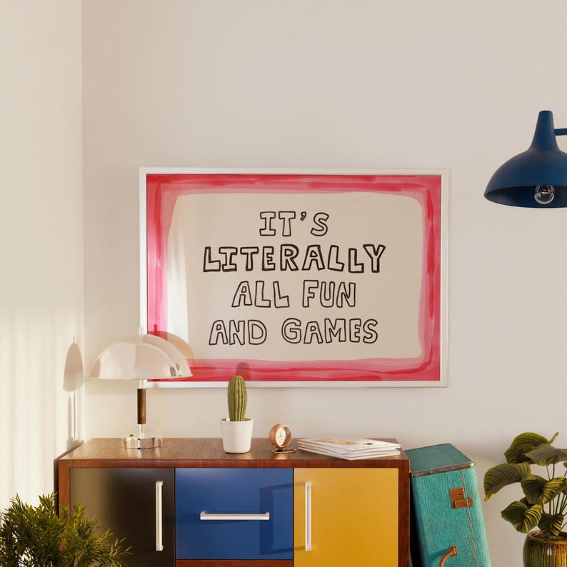 May include: A rectangular wall art print featuring the black text IT'S LITERALLY ALL FUN AND GAMES centered on a cream background, enclosed by a vibrant pink painted border. The print is displayed in a thin white frame, hanging on a plain wall.