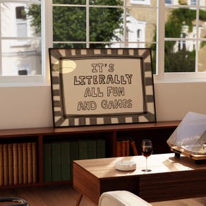 May include: A rectangular framed wall art piece featuring a beige background with a dark gray striped border. The text "IT'S LITERALLY ALL FUN AND GAMES" is printed in the center in a bold, hand-drawn style black font with a casual, uneven appearance.