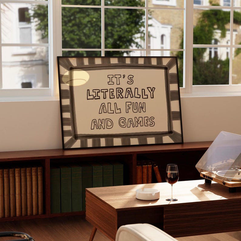 May include: A rectangular framed wall art piece featuring a beige background with a dark gray striped border. The text "IT'S LITERALLY ALL FUN AND GAMES" is printed in the center in a bold, hand-drawn style black font with a casual, uneven appearance.