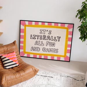 May include: A rectangular framed wall art piece featuring a white background with a yellow border and a pink and white striped sunburst pattern. The text IT'S LITERALLY ALL FUN AND GAMES is printed in black, hand-drawn style capital letters.