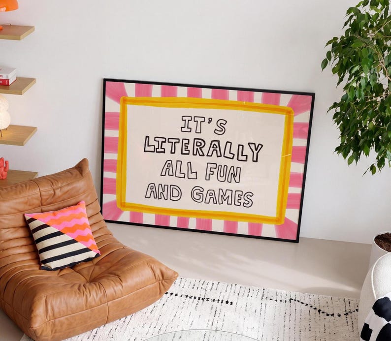 May include: A rectangular framed wall art piece featuring a white background with a yellow border and a pink and white striped sunburst pattern. The text IT'S LITERALLY ALL FUN AND GAMES is printed in black, hand-drawn style capital letters.