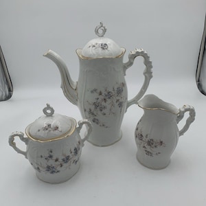 Könnte beinhalten: Ein weißes Porzellan-Teeservice mit Teekanne, Zuckerdose und Sahnekännchen. Das Set hat ein blau-violettes Blumenmuster mit goldenen Verzierungen. Die Teekanne ist etwa 25 cm hoch.