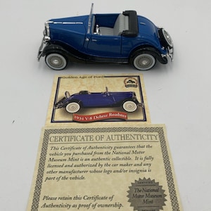 Puede incluir: Un coche modelo azul 1934 V-8 Deluxe Roadster con neumáticos de banda blanca. El modelo se encuentra encima de un certificado de autenticidad del National Motor Museum Mint. El certificado indica que el coche es un objeto de colección auténtico.