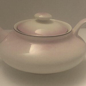 May include: A white teapot with a pink and white ombre glaze. The teapot has a lid and a handle.