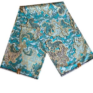 Op de afbeelding: Een gevouwen stuk stof met een turquoise en gouden barokpatroon. De stof heeft wervelende bloemmotieven in tinten turquoise, goud en crème. De stof is waarschijnlijk katoen of een vergelijkbaar materiaal, geschikt voor kleding of woondecoratie.