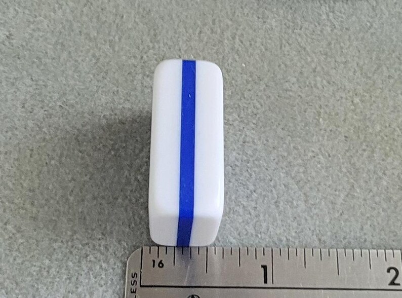 May include: A white rectangular object with a vertical blue stripe down the center. The object is next to a ruler, and appears to be approximately 1 inch in length.